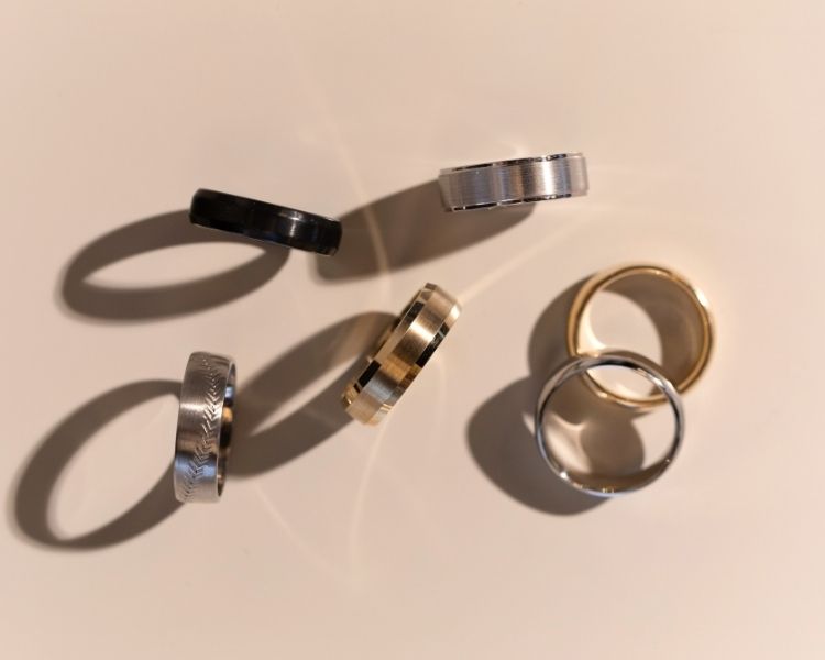 A curated selection of TOVAA men's wedding bands in gold, platinum, tungsten, and black finishes, arranged in soft light to highlight texture and form.